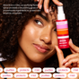 A woman with curly hair smiles, holding Shielded Beauty Face the Day Purifying Mist by her face. Text highlights its nourishing facial spray formula that shields skin with antioxidants, providing purifying and protective benefits.