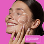 A woman applies Shielded Beauty’s Triple Threat Treatment patch under her eye. Text highlights clean beauty with hyaluronic acid and vitamin B12 for hydration, priming, masking, and brightening. Background is bright pink.