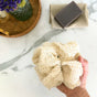 A hand holds the Me Mother Earth Reusable Sisal Shower Pouf in beige, eco-friendly and carefully knitted, above a marble surface. Nearby, a bar of soap rests on a textured cloth beside a basket featuring a jar filled with purple flowers.