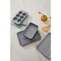The Caraway Mini Bakeware Set W/ Storage by Caraway, featuring a muffin pan, baking tray with cooling rack, and loaf pan, is displayed on a marble countertop with a wooden spatula, pastel oven mitts, and pink tulips in a vase.