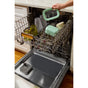 An open dishwasher loaded with dishes and a green Caraway Mini Food Storage Set. A hand places the matching lid on the top rack. The organized kitchen features wooden floors and a white countertop.