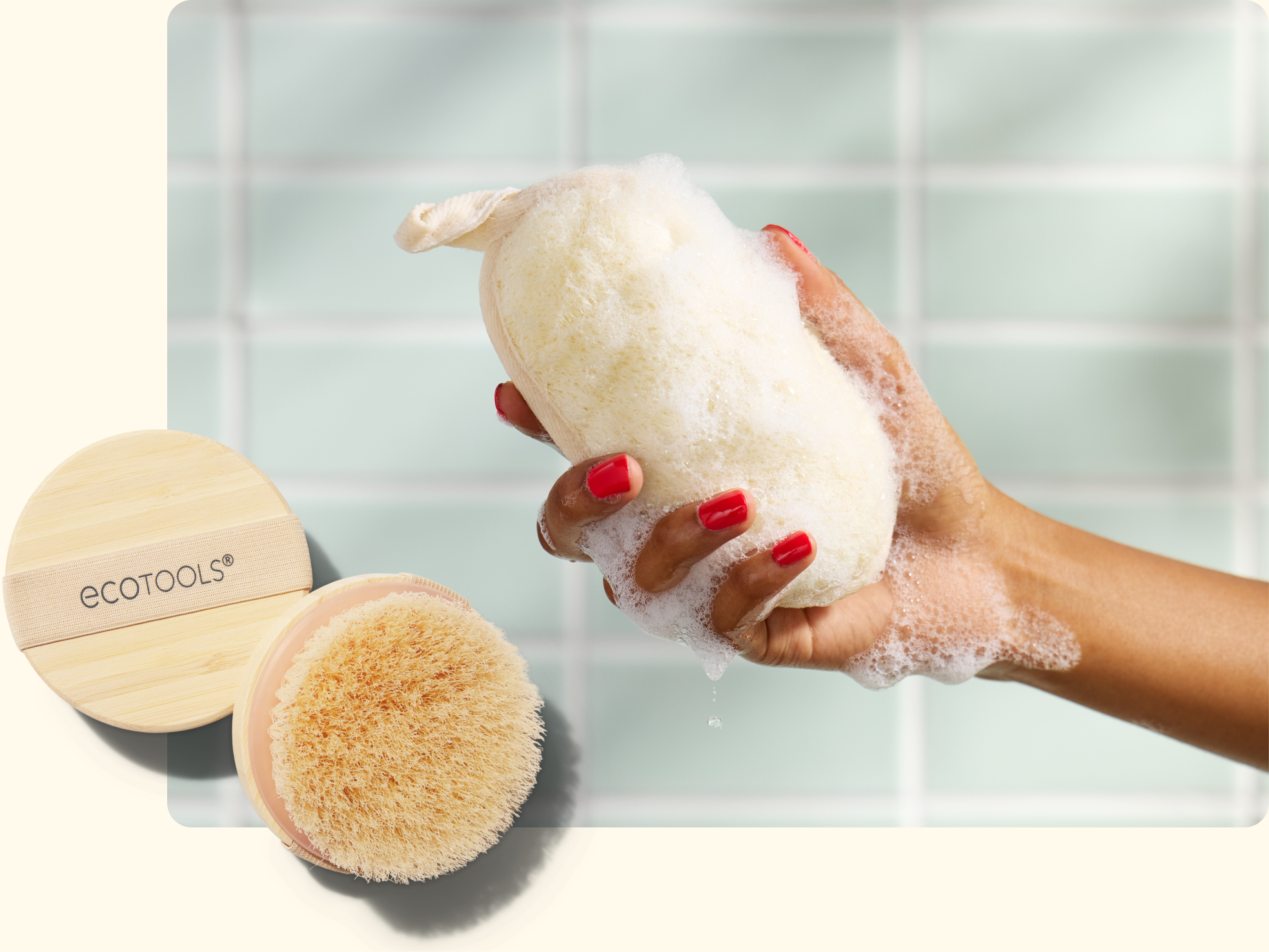 A hand with red nail polish holding a soapy bath sponge, with a wooden EcoTools body brush placed nearby. The background features light green tiled walls.