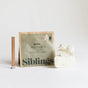 A Siblings DIY Candle Kit is shown: a green refill pouch stands upright beside a wooden wick and three pieces of white wax, all against a plain white background—ideal for eco-friendly candle making with compostable packaging.