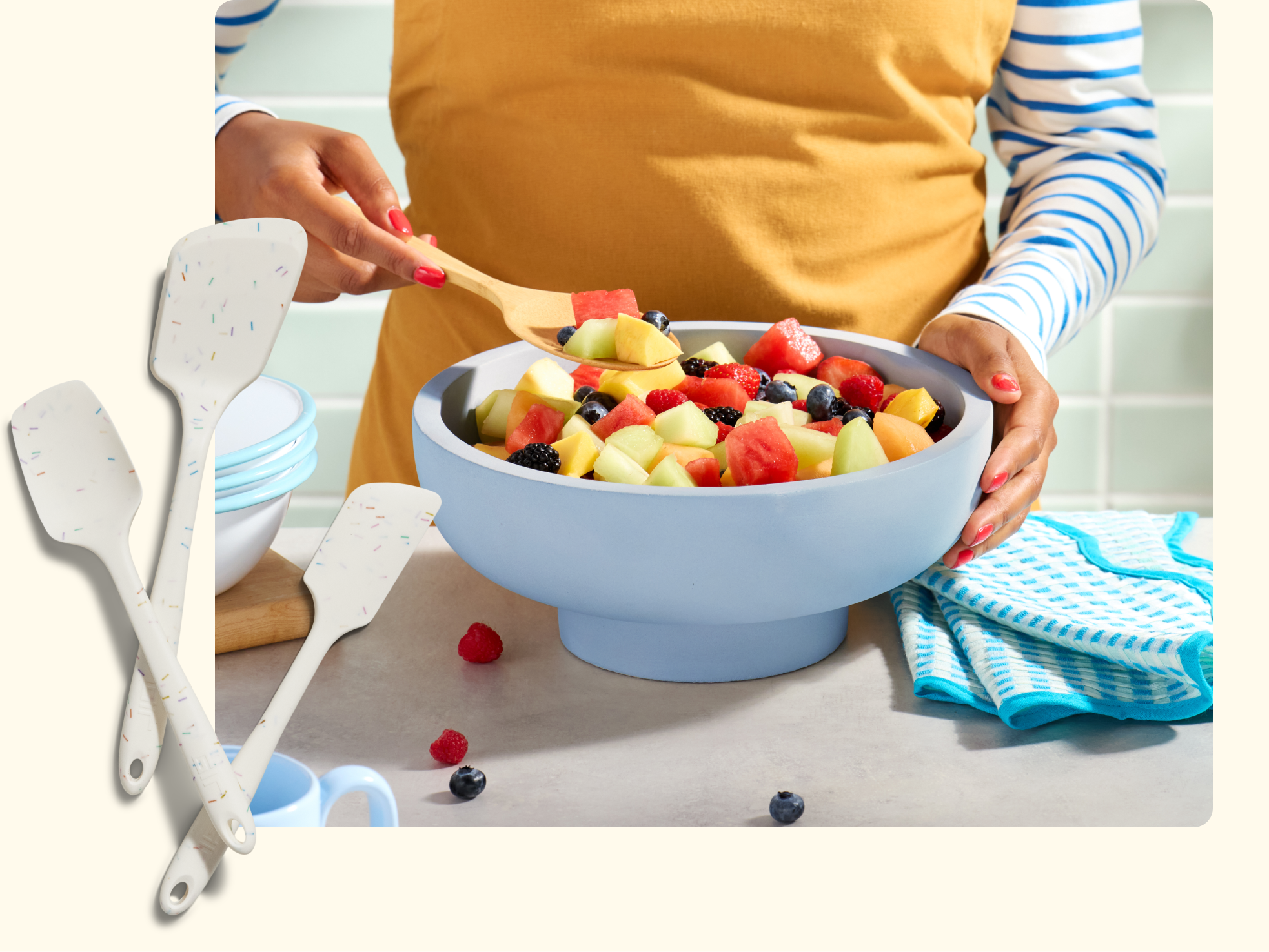 A person in a yellow apron mixes a colorful fruit salad in a light blue bowl, surrounded by kitchen towels, a mug, and three white spatulas with speckled handles on a light counter.