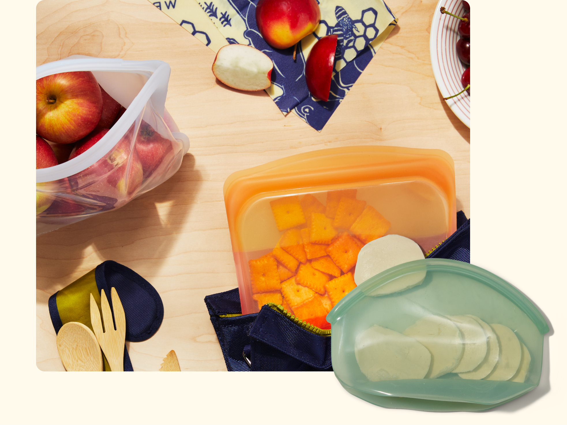 Reusable silicone bags filled with sliced apples, cheese cubes, and cucumber slices sit on a wooden table with a blue napkin, wooden utensils, and pieces of fruit.