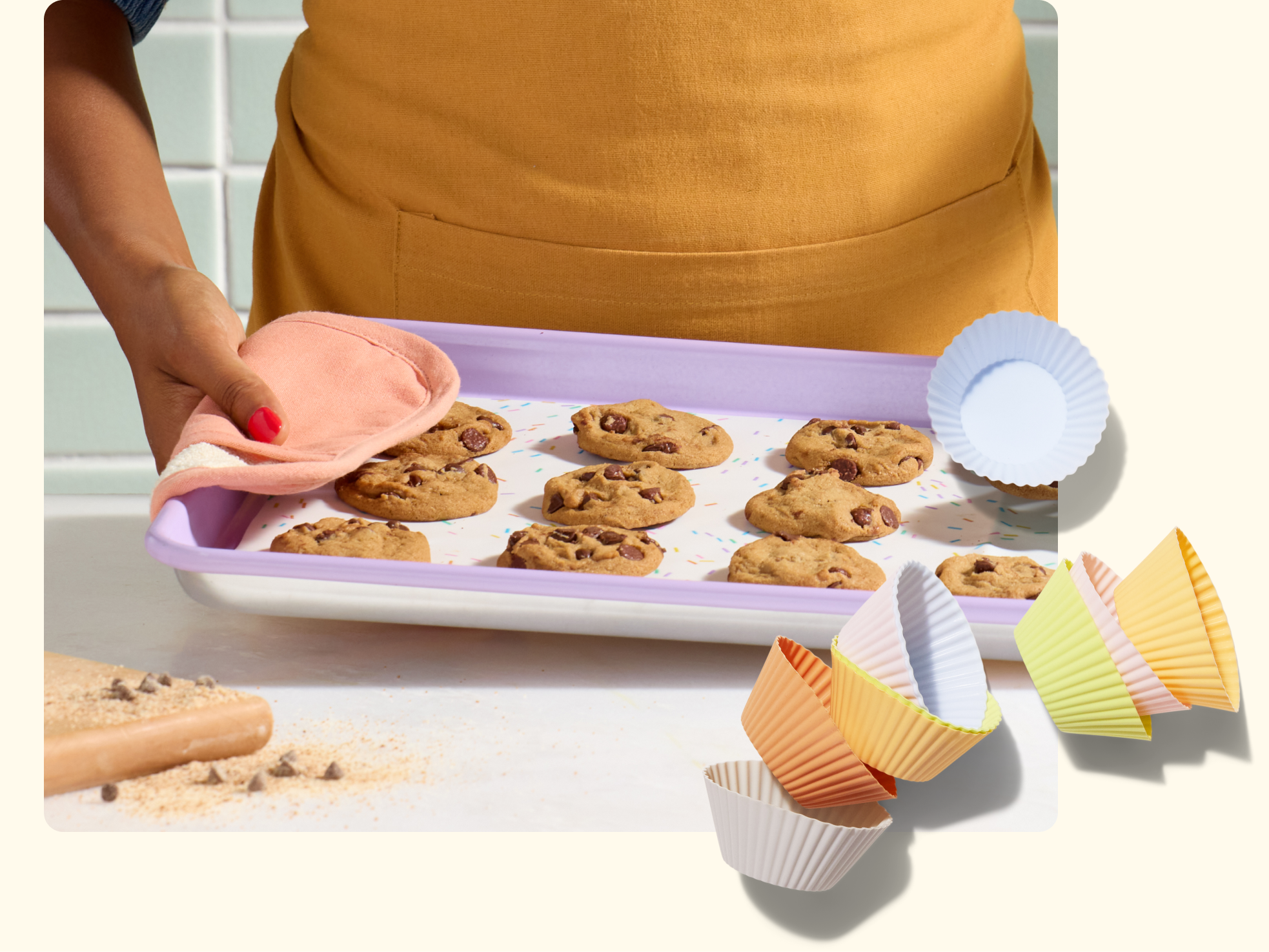 A person in a yellow apron holds a tray of freshly baked chocolate chip cookies with a pink oven mitt. Colorful cupcake liners are scattered in the foreground.