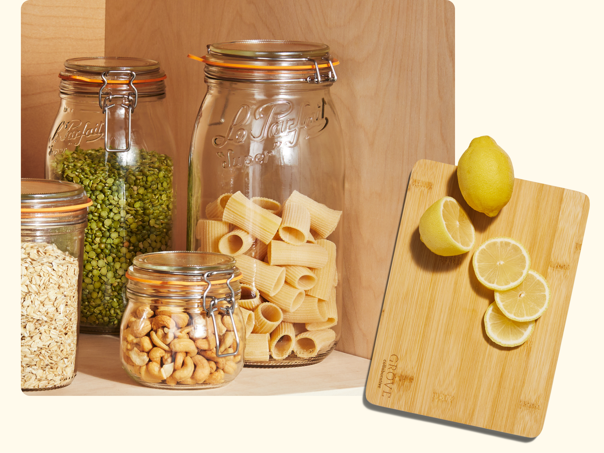 Four glass jars containing green split peas, rigatoni pasta, oats, and cashews are grouped together on a wooden surface. Next to them, a wooden cutting board holds a whole lemon and lemon slices.