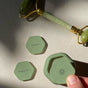 A hand holds a green hexagonal Cadence capsule with a sun symbol. Nearby are two matching Cadence Labels - Set of 6 lids marked Cleanser and Moisturizer. A jade roller rests above on a neutral surface as sunlight creates soft shadows.