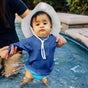 A baby in a blue Green Sprouts Eco Snap Swim Diaper, made from recycled polyester (24 months, 24-28 lbs), wears a white sun hat while standing in a shallow pool with an adults gentle support. The focused child watches ripples as the adult holds their arms.
