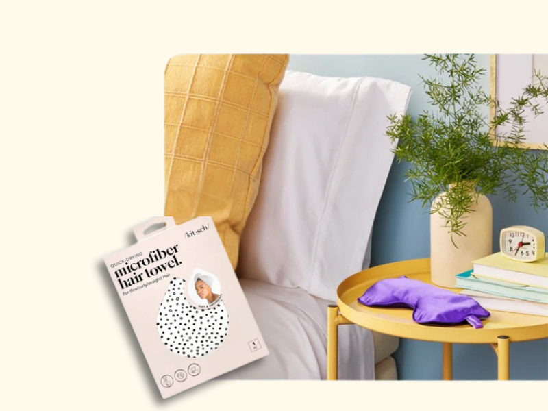 A bed with white sheets and a yellow pillow is next to a round yellow side table holding a clock, books, a plant, and a purple eye mask. In the foreground is a box for a microfiber hair towel.
