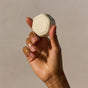 A hand holds a small, beige, hexagonal Cadence Hair Duo Travel Container with rounded edges against a plain, neutral background.