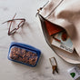 A beige handbag, eyeglasses, keys, mints, and a blue Stasher Reusable Silicone Sandwich Bag with pretzels rest on marble. Partially inside the bag are a brown wallet and a magazine.