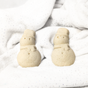 Two small Spinster Sisters Co. Holiday Bath Bombs - Snowman, made of beige dough, are side by side on a soft white speckled blanket with a plush, cozy background.