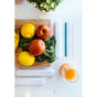 A wooden board with apples, lemons, and kale sits on a white table next to the Full Circle Soma Glass Travel Straw in teal, its straw case, a glass of orange juice, and a small potted plant.