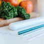A close-up of the Full Circle Soma Glass Travel Straw - Teal on a countertop with leafy kale and apples in the background, highlighting a healthy, eco-friendly lifestyle.