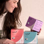 A smiling woman holds a purple box of Sparkle Maxi Pads, With Wings | 10 Count, while two more boxes—one pink and one blue—sit on the table in front of her, each featuring the Sparkle brand and product details.
