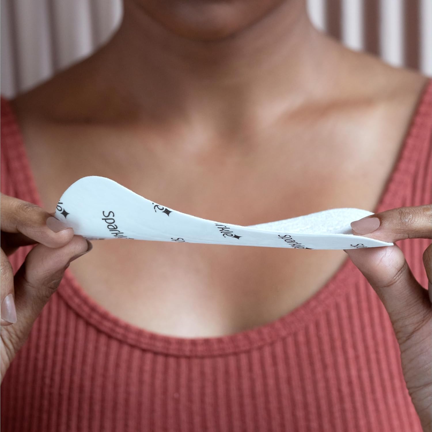 A person in a pink ribbed tank top holds a Sparkle Panty Liner—a hypoallergenic, flexible, curved adhesive pad with printed text—horizontally between their hands.
