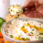 A hand holds a cucumber slice with creamy dip, garnished with herbs and Kosterina Spicy Red Pepper Olive Oil. Below, a bowl of the dip is drizzled with Kosterina’s olive oil and surrounded by fresh veggies.