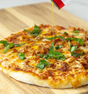 A close-up of a pizza with melted cheese, meat, and basil on a wooden board, as Kosterina Spicy Red Pepper Olive Oil is drizzled over it from the can.