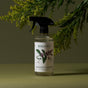 A bottle of Koala Eco Stainless Steel Cleaner with a black spray top and botanical label sits on a green surface, ideal for cleaning stainless steel, with leafy branches in the background.