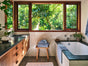 Sunlit bathroom with a large window overlooking lush green trees, a soaking tub draped with an absorbent Coyuchi Air Weight® Organic Towel, wooden cabinets, a bench with folded Coyuchi Air Weight® Organic Towels, and white subway tile walls.