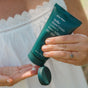 A person in a white, lace-trimmed top squeezes Ursa Major’s Stellar Shave Cream from a green tube onto their palm, wearing a gold ring set with a light blue stone.