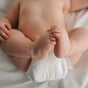 A close-up of a baby lying on a white sheet, legs crossed and feet visible, perfectly showing gentle care with ATTITUDE baby leaves Diaper Cream for zinc oxide diaper rash relief.