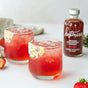Two glasses of a pink-red iced drink with sugared rims sit on a white surface. Nearby is a bottle of Kosterina Crushed Strawberry Vinegar, with fresh strawberries and a napkin—perfect for fruit salads without added sugar.