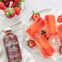 A plate with four strawberry popsicles on ice, surrounded by fresh strawberries, a bowl of fruit salad, and a bottle of Kosterina Crushed Strawberry Vinegar on a white surface with a textured towel—perfect summer treats with no added sugar.