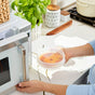 A person holds a round food container sealed with a Stasher Reusable Stretch Lid (Small, 3-pack), preparing to place it in the microwave on a kitchen counter with plants and kitchen items in the background.
