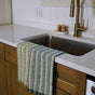A Supra Endura Striped Organic Cotton Tea Towel hangs over a stainless steel sink with gold faucet, set in a white countertop above wooden cabinets. Two scrub brushes rest on a plate near the faucet.