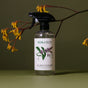 A bottle of Koala Eco Natural Glass Cleaner with a black spray nozzle sits before a green background and yellow flowers. This cleaner leaves windows sparkling and smear-free.