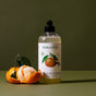 A bottle of Koala Eco Natural Fruit & Vege Wash, a natural cleaner, is on a green surface next to a peeled mandarin with leaves, set against an olive green background.