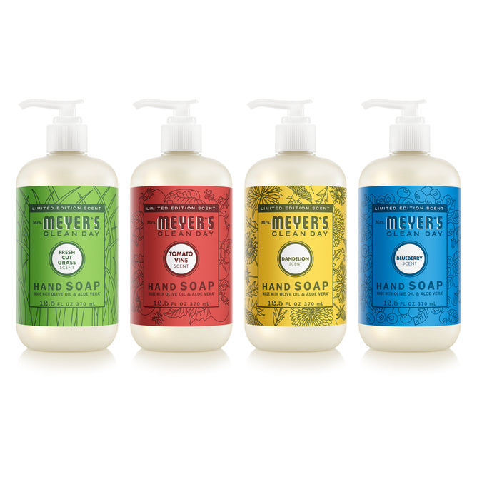 Four bottles from the Mrs. Meyer's Summer Hand Soap Collection are lined up, each with a colorful label: Fresh Cut Grass (green), Tomato Vine (red), Dandelion (yellow), and Blueberry (blue).