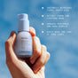 A hand holds a bottle of Ursa Major Sunny Start Eye Serum up to a blue sky, with text calling out its benefits: this eye serum brightens and hydrates the under-eye area, helps firm skin, visibly reduces puffiness and dark circles, and smooths the look of fine lines.