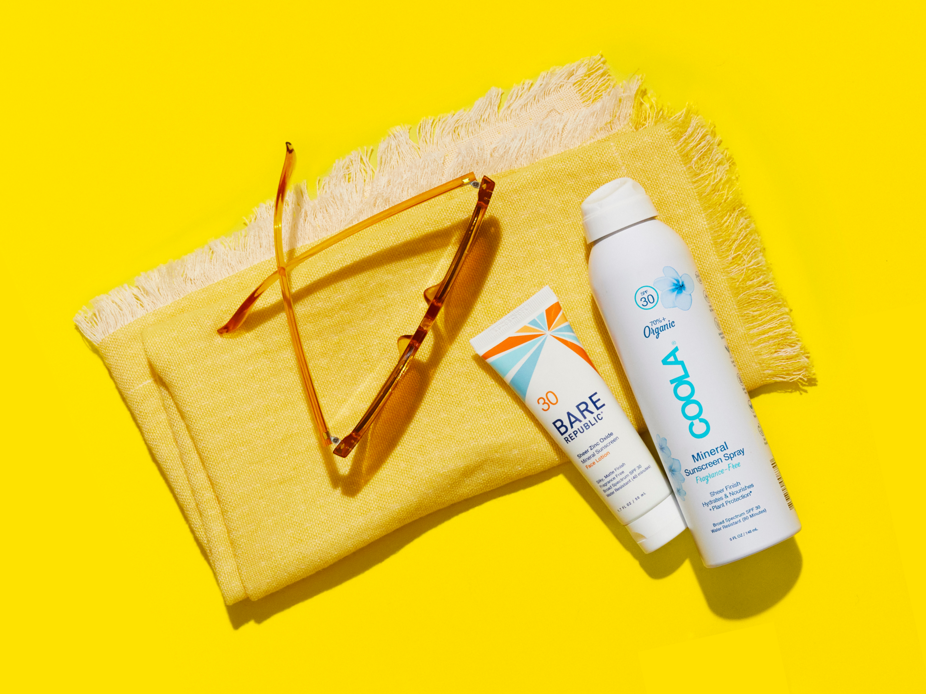 Sunscreen, spray sunscreen, yellow sunglasses, and a yellow towel are arranged on a bright yellow background.
