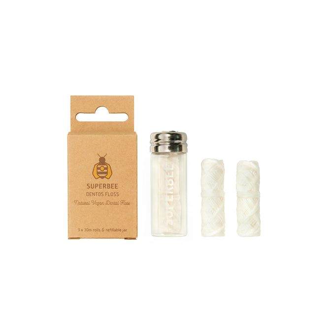A SuperBee Dentos Eco Dental Floss box sits next to a small glass jar with a metal lid containing plastic-free vegan dental floss, along with two spare white floss rolls standing upright.