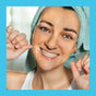 A smiling woman in a white tank top and gold hoop earrings flosses with SuperBee Dentos Eco Dental Floss, her head wrapped in a towel. The image is framed by a light blue border.