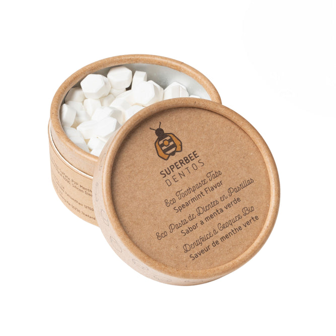 An open round SuperBee container labeled "Dentos Eco Toothpaste Tabs, Spearmint Flavor" shows white hexagonal tabs. The vegan, eco-friendly tabs offer refreshing spearmint taste in a travel-friendly format.
