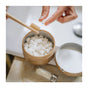 A person holds a Dentos Eco Toothpaste Tab and a bamboo toothbrush over an open container of travel-friendly Spearmint tabs by SuperBee, while a small glass SuperBee bottle rests on the bathroom counter.