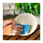 A person washes a white plate with soapy water using the SuperBee Kitchen Loofah Dish Sponge over a wooden surface, with blurred green plants in the background.