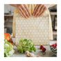 A person in a yellow dress holds a SuperBee Waxed Food Bag with a hexagon pattern. Fresh produce like lettuce, strawberries, ginger, and papaya is arranged on the counter beside their SuperBee beeswax bags.