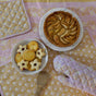A top view of a table set with the Supra Endura Hand Block Printed Ovenmitt And Pot Holder Set, a wooden spoon, an apple tart on a white plate, and a bowl of assorted cookies on a pastel-patterned tablecloth.