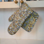The Hand Block Printed Ovenmitt and Pot Holder Set by Supra Endura features a floral pattern in yellow, white, and grey, shown hanging from kitchen cabinet knobs.