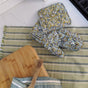 The Supra Endura Hand Block Printed Ovenmitt And Pot Holder Set rests on a green striped kitchen towel next to a wooden cutting board and spoon on a white countertop near the stove.