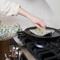 Wearing the Supra Endura Hand Block Printed Ovenmitt and using the matching Pot Holder, a person lifts a hot pan lid placed on a lit gas stove burner.