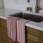 A Supra Endura Striped Organic Cotton Tea Towel hangs over a white marble counter by a stainless steel sink with a brass faucet. Wooden cabinets and spice jars are seen in the background.