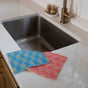 Two Supra Endura Swedish Dish Cloths—one blue, one pink—are displayed on a marble kitchen countertop beside a stainless steel sink and gold faucet, offering super absorbency for eco-friendly cleaning.