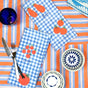 A table setting with a blue-orange striped cloth and blue-checked napkins featuring tomatoes and carrots is ideal for tomato lovers. A patterned bowl, plate, blue glass, and silverware on the Tomato Print Organic Cotton Kitchen Towel by Supra Endura complete the look.
