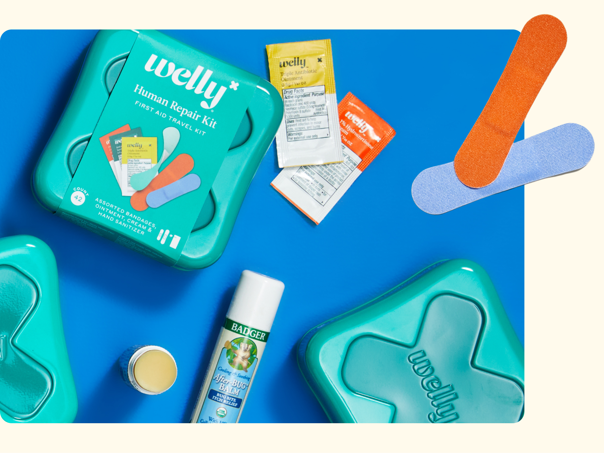 Flat lay of Welly Human Repair Kit with teal tins, assorted bandages, ointment packets, a can of bug spray, and a lip balm stick on a blue background. Two bandages are displayed separately.